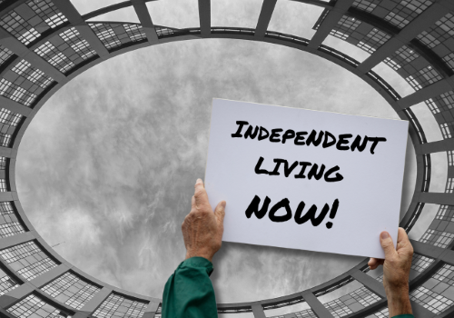 Le braccia di una persona sollevano un cartello su scritto: “Independent Living Now!”, ovvero “Vita Indipendente ora!”