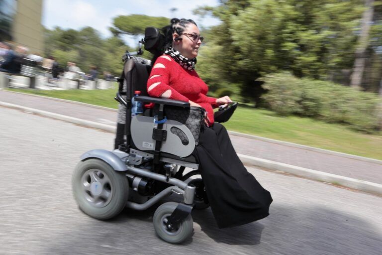 Una donna con disabilità sfreccia con la sua sedia a rotelle lungo una strada in mezzo al verde.