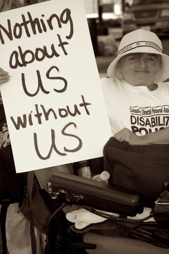 Foto in bianco nero di una donna con disabilità che manifesta per i propri diritti esibendo un cartello con la scritta “Nothing about us without us” (Nulla su di noi senza di noi). Lei potrebbe essere un bell’esempio di “Donna Convenzione”.
