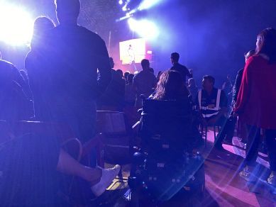 Una persona in sedia a rotelle, ritratta di spalle, segue una partita su dei maxischermi assieme ad un gruppo di altre persone.