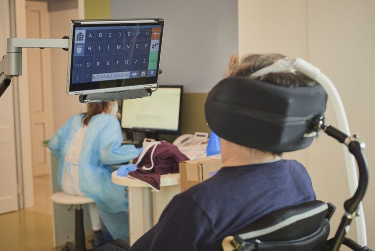 Un uomo con sclerosi laterale amiotrofica ritratto di spalle presso uno dei Centri Clinici Nemo, mentre, sullo sfondo, un’operatrice sanitaria utilizza un computer.