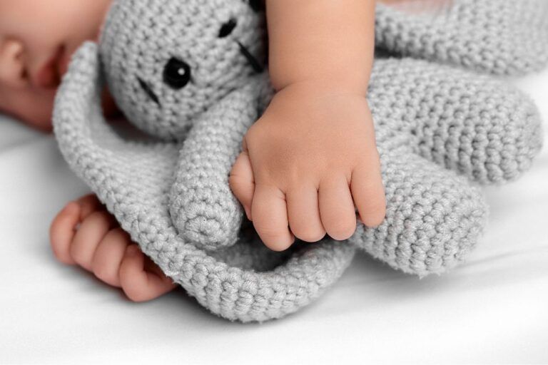 Particolare di un neonato che dorme tenendo stretto tra le braccia un pupazzo grigio.